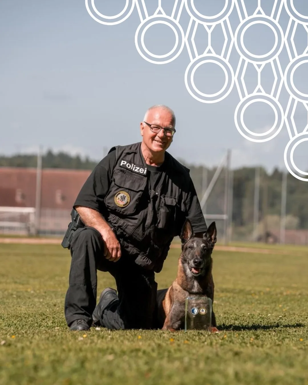 Ein Polizist in schwarzer Uniform kniet auf einer Wiese neben einem belgischen Schäferhund. Im Hintergrund sind Bäume sichtbar.