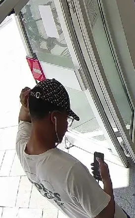 Ein Mann mit Kappe und Ohrhörern steht vor einer Glastür und blickt auf sein Smartphone. Er trägt ein weißes T-Shirt.