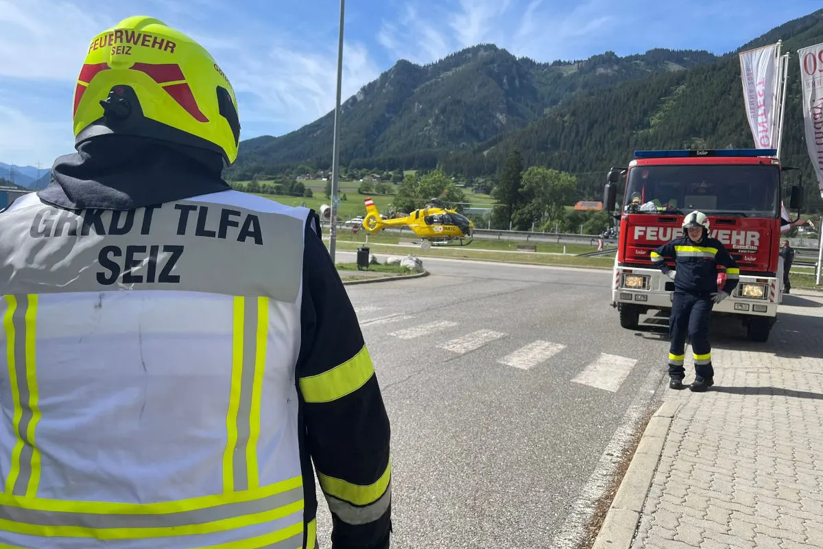 Ein Feuerwehrmann steht auf einer Straße zugewandt, ein Feuerwehrauto und ein Rettungshubschrauber sind ebenfalls zu sehen. Berge im Hintergrund.