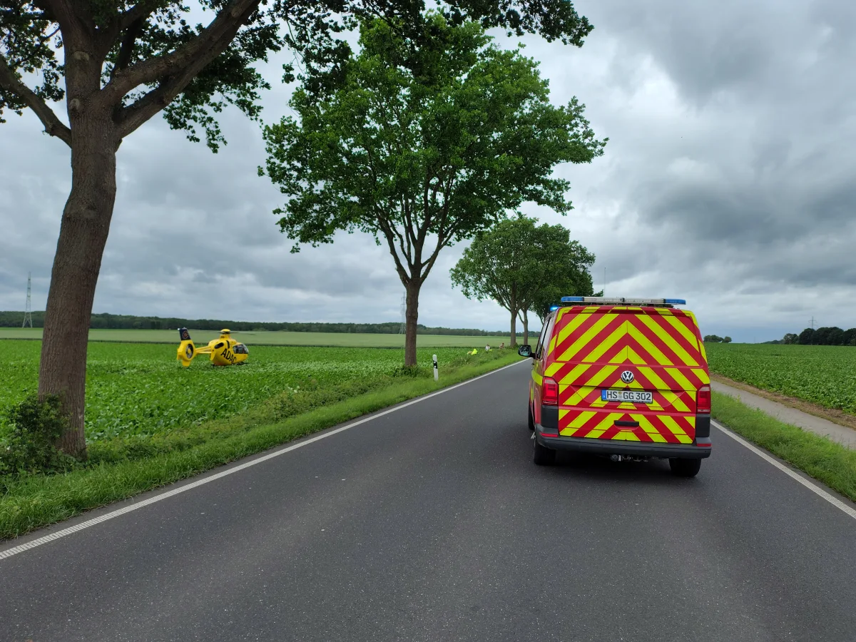 Ein Rettungswagen ist auf einer Landstraße geparkt, während ein gelber ADAC-Hubschrauber in einem Feld dahinter sichtbar ist. Die Szene zeigt eine ländliche Umgebung mit Bäumen und bewölktem Himmel.