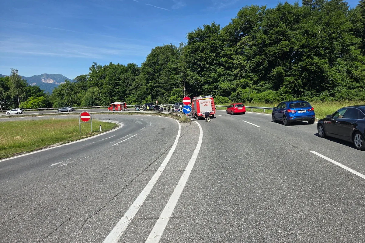 Mehrere Fahrzeuge, darunter ein Feuerwehrwagen, befinden sich auf einer kurvigen Landstraße umgeben von Bäumen bei sonnigem Himmel.
