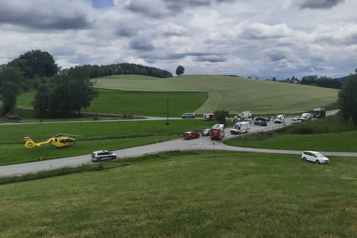 Mehrere Einsatzfahrzeuge und ein Rettungshubschrauber sind an einer Landkreuzung positioniert. Die Szene zeigt grüne Felder und bewölkten Himmel.
