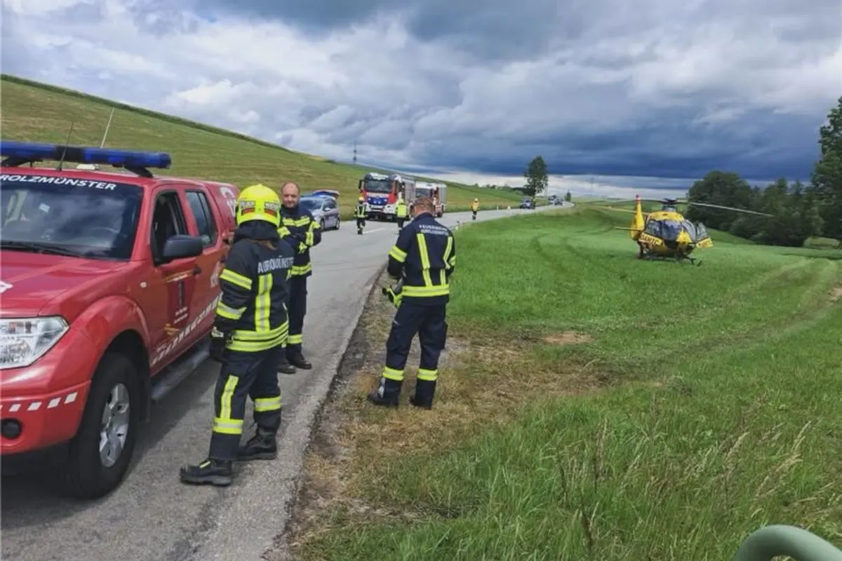 Feuerwehrleute stehen neben einem roten Einsatzfahrzeug auf einer Landstraße. Ein gelber Rettungshubschrauber steht auf der Wiese bereit.