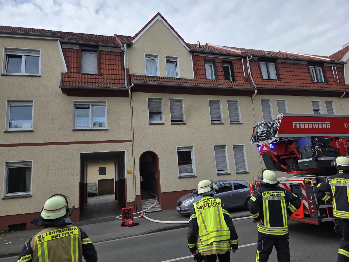 Mehrere Feuerwehrleute stehen vor einem Wohnhaus, ein Feuerwehrfahrzeug ist vor Ort im Einsatz.
