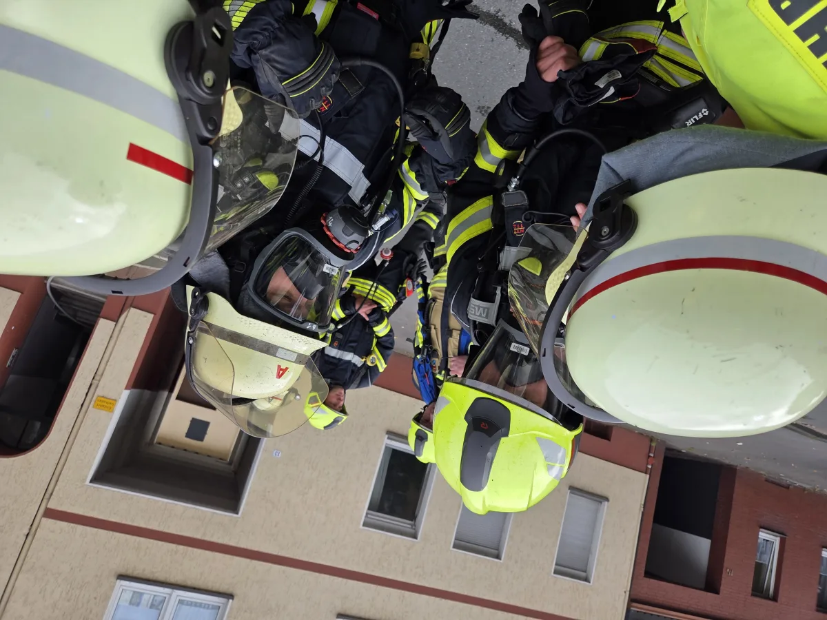 Mehrere Feuerwehrleute in gelben Schutzanzügen und Helmen stehen dicht beieinander. Hinter ihnen ist ein großes Gebäude zu sehen.