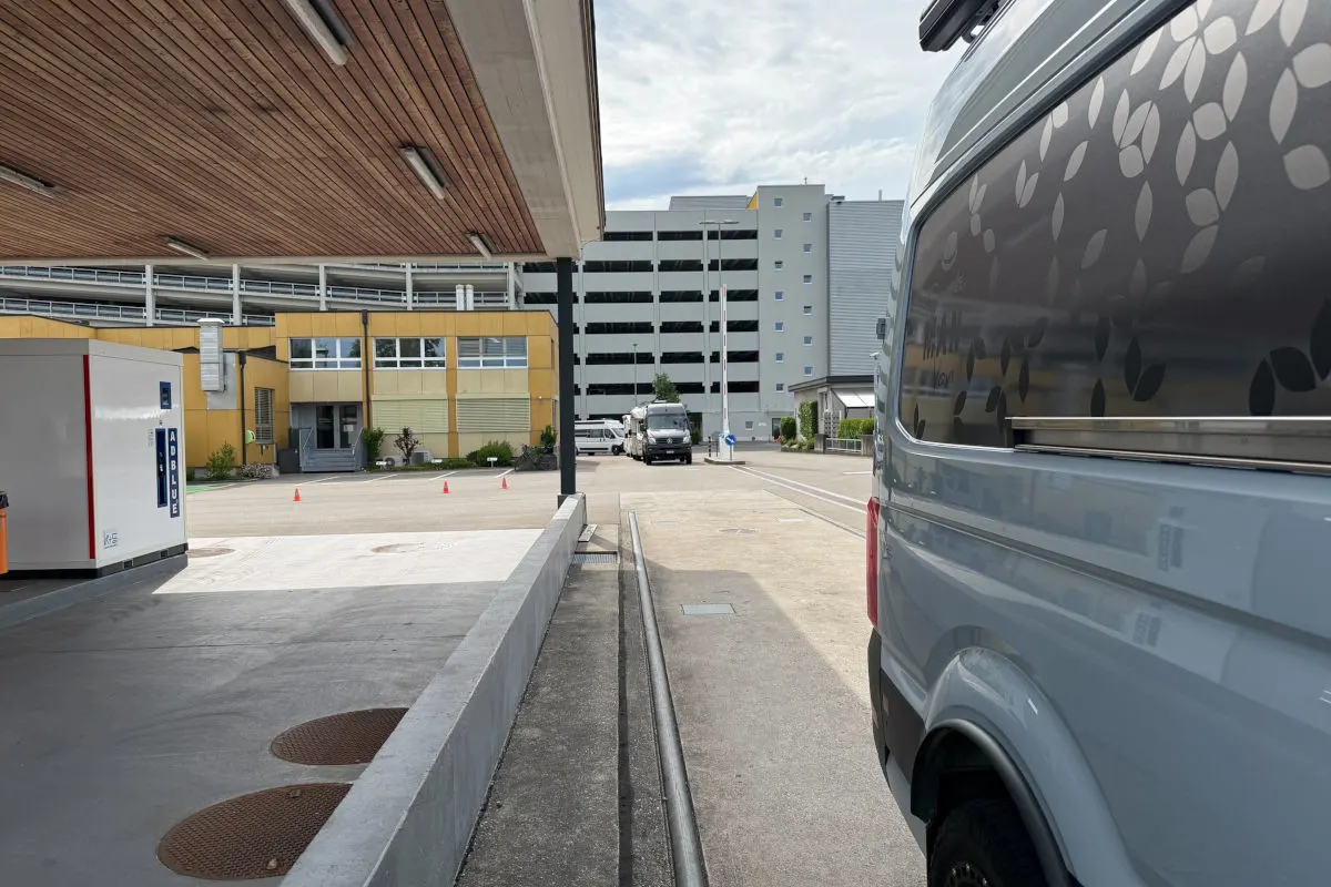 Ein Transporter parkt auf einem asphaltierten Parkplatz neben einem gelben Verwaltungsgebäude. Im Hintergrund sind weitere Gebäude sichtbar.
