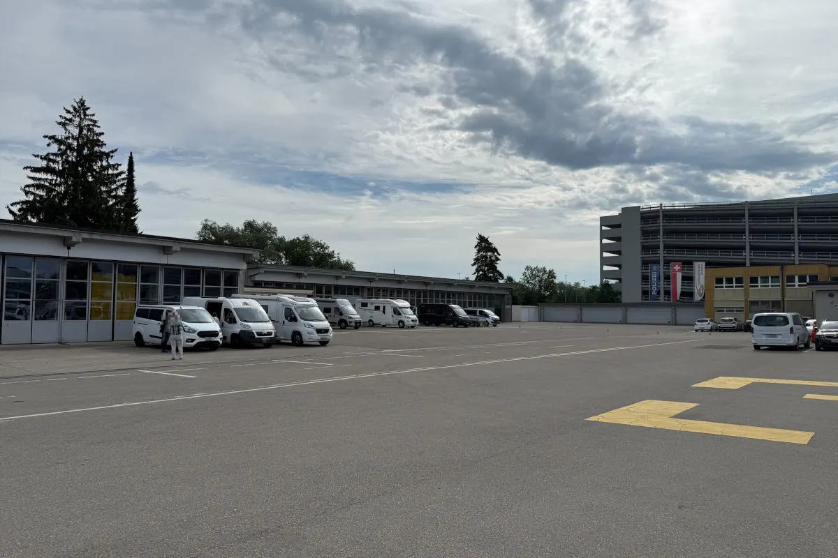 Ein großer Parkplatz mit mehreren weißen Transportern, die vor einem Gebäude geparkt sind. Im Hintergrund ist ein mehrstöckiges Gebäude sichtbar, und der Himmel ist teilweise bewölkt.