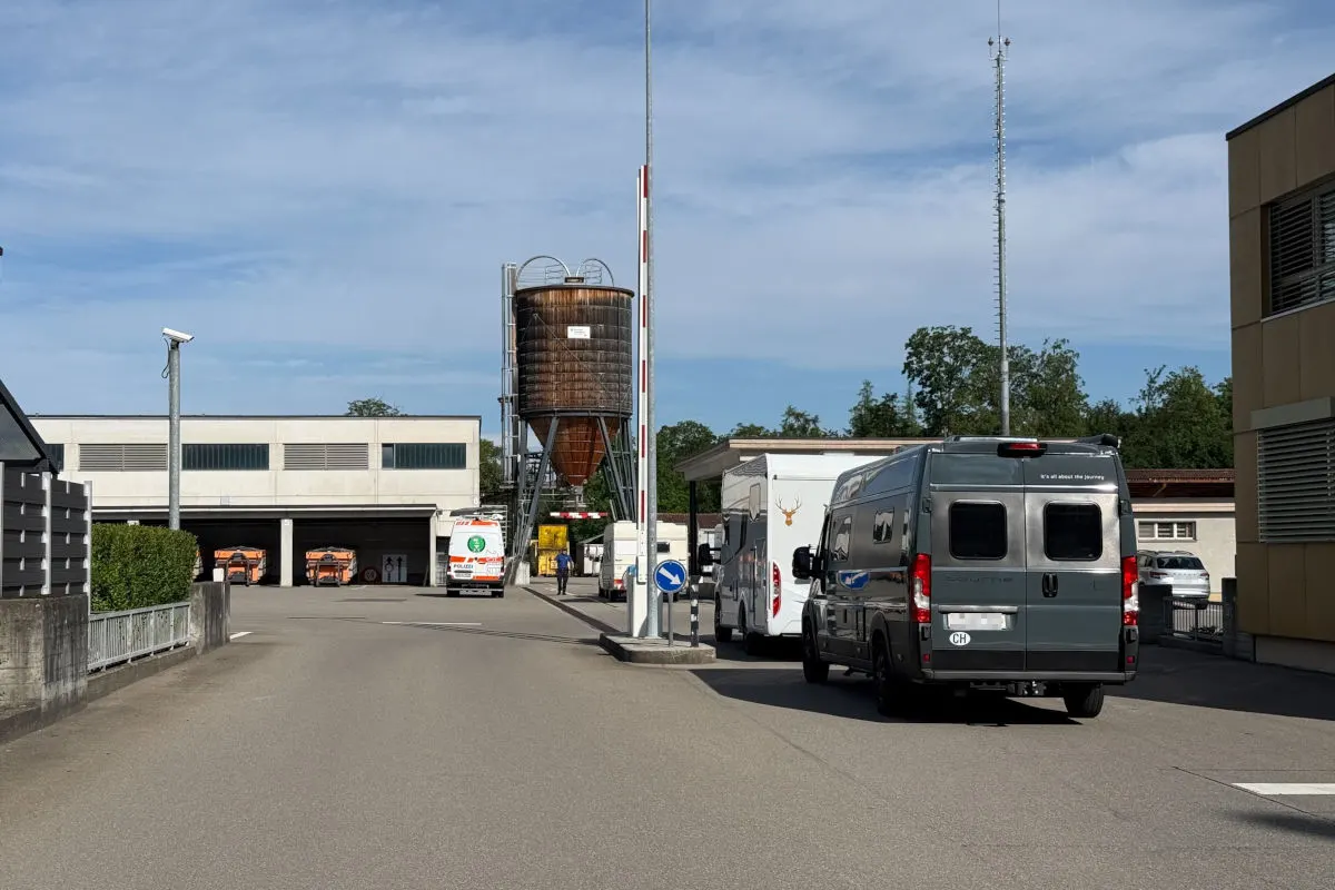 Ein Siloturm steht in einem Industriegebiet, umgeben von mehreren geparkten Fahrzeugen. Die Szene wirkt ruhig und ist bei Tageslicht aufgenommen.