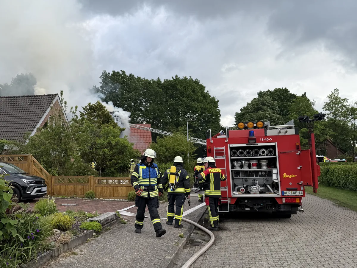 Feuerwehrleute stehen neben einem Löschfahrzeug, während Rauch aus einem Hausdach austritt. Der Himmel ist bewölkt.