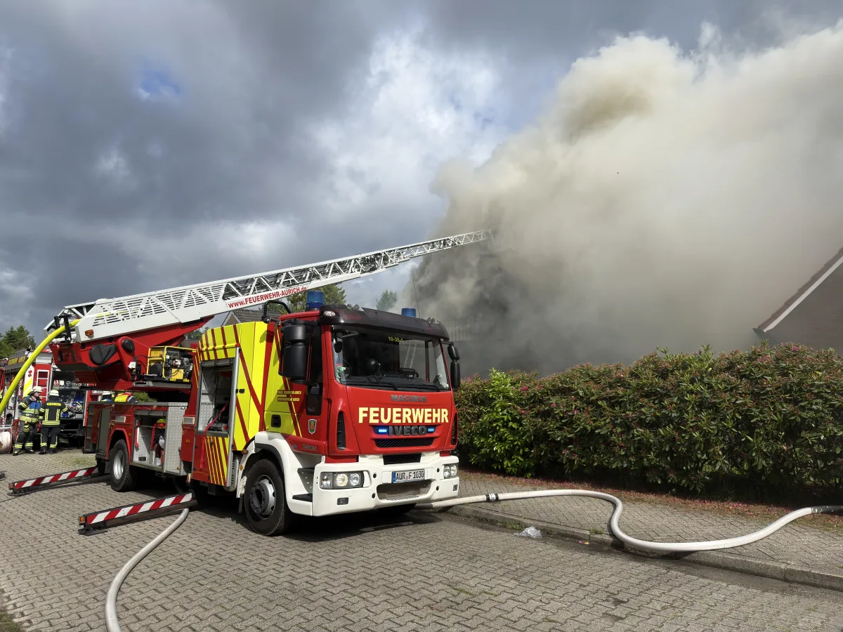 Ein rotes Feuerwehrfahrzeug mit ausfahrender Leiter steht vor einem Gebäude, aus dem dichter Rauch aufsteigt. Zwei Feuerwehrleute sind im Einsatz.