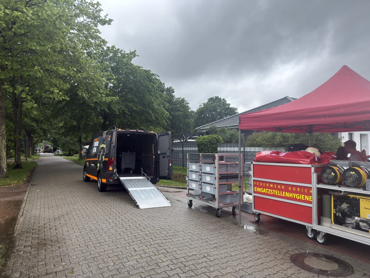 Ein schwarzer Van mit offener Heckklappe steht auf einer Wohnstraße. Rechts daneben ist ein rotes Zelt mit Einsatzmaterialien der Feuerwehr Aurich zu sehen.