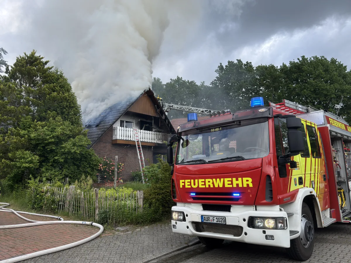 Ein roter Feuerwehrwagen steht vor einem Haus, dessen Dach in Flammen steht und dichte Rauchwolken aufsteigen. Das Haus befindet sich in einer grünen Umgebung.