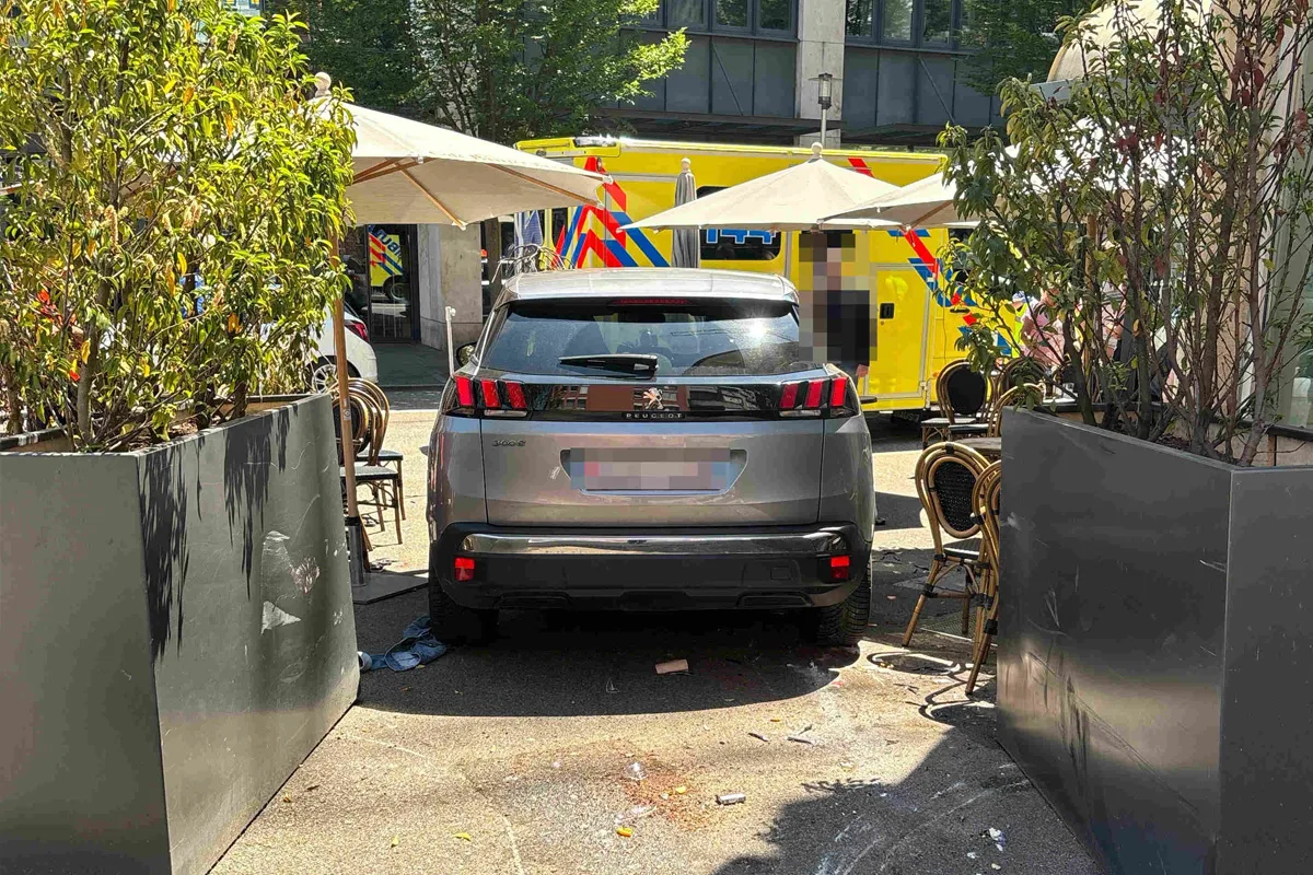 Ein grauer SUV parkt in einem Außenbereich eines Cafés, umgeben von Stühlen und Sonnenschirmen, mit einem gelben Krankenwagen im Hintergrund.