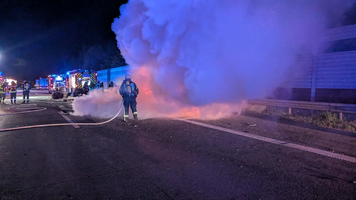 Ein Feuerwehrmann bekämpft einen Brand auf einer nächtlichen Autobahn mit Schläuchen, während Rauch aufsteigt. Mehrere Einsatzfahrzeuge sind im Hintergrund zu sehen.