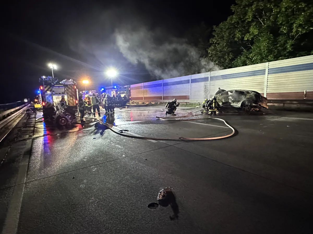 Feuerwehrleute sind auf einer abendlichen Autobahn zu sehen, wie sie einen ausgebrannten Pkw löschen. Im Hintergrund sind Feuerwehrfahrzeuge mit eingeschaltetem Blaulicht.