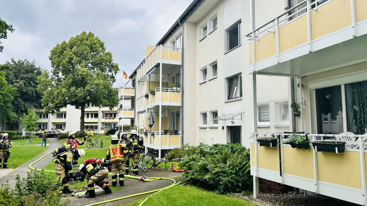Mehrere Feuerwehrleute in voller Ausrüstung stehen vor einem mehrstöckigen Wohngebäude mit Balkonen. Auf dem Boden liegen Schläuche und Ausrüstungsteile.
