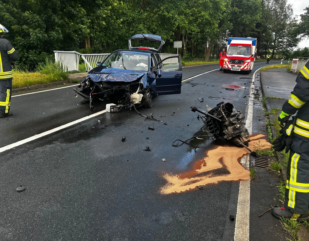 Ein stark beschädigtes Auto steht auf einer nassen Straße; daneben Feuerwehrleute und Trümmer. Ein Rettungsfahrzeug ist ebenfalls zu sehen.