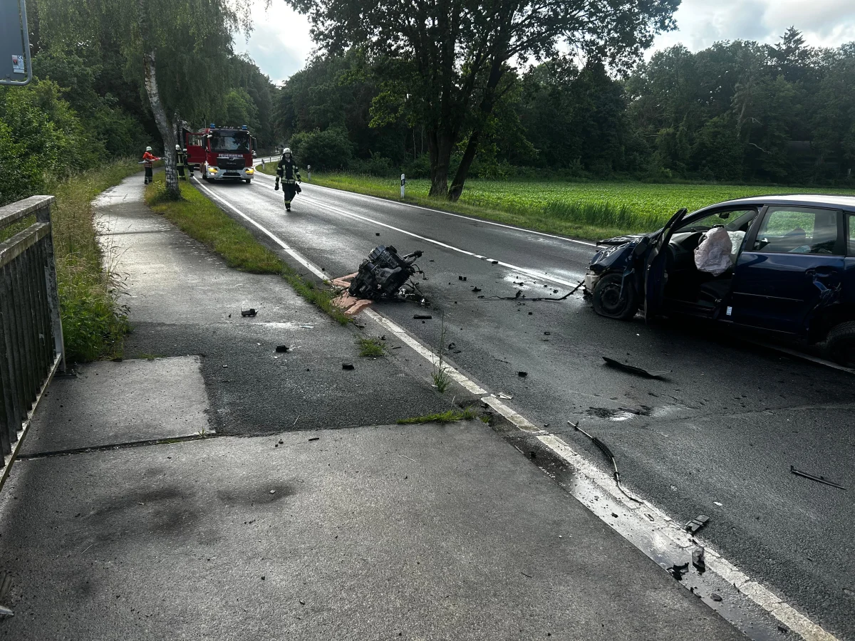 Auf einer Landstraße steht ein stark beschädigtes blaues Auto mit geöffneten Türen. Trümmer sind auf der Straße verteilt und ein Feuerwehrfahrzeug ist im Hintergrund zu sehen.