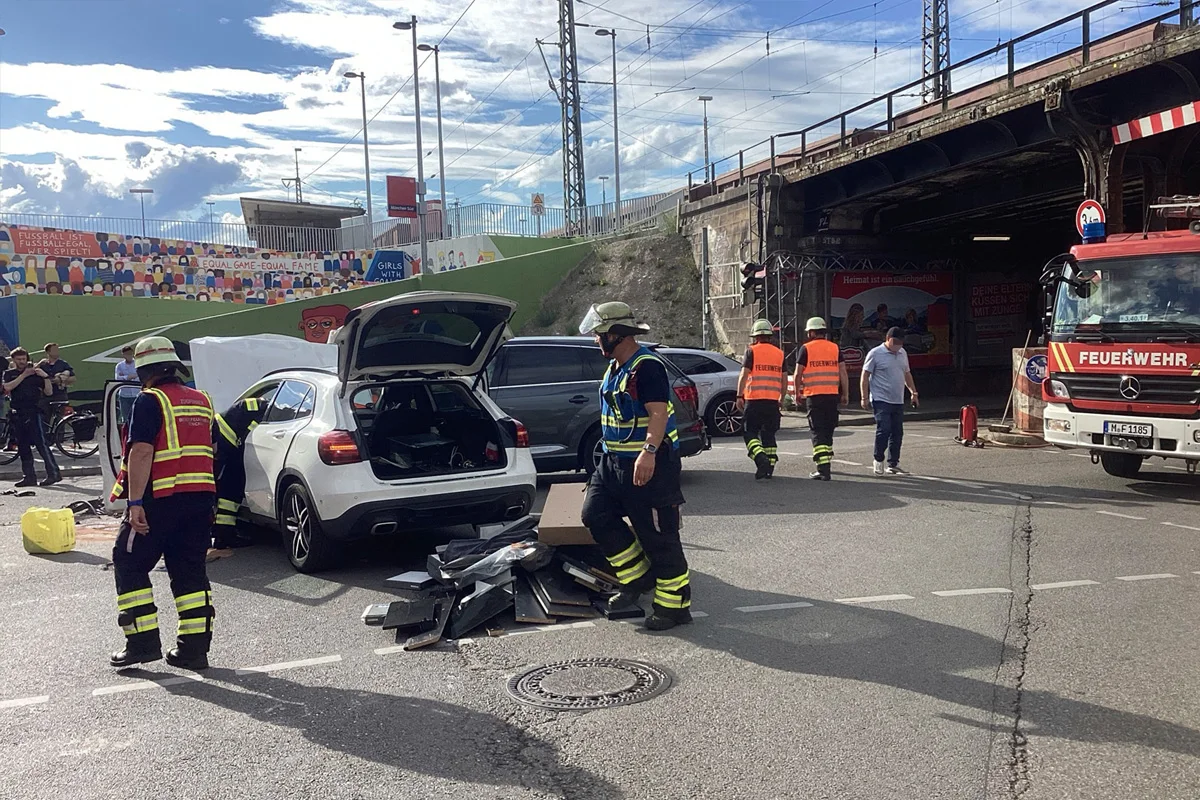 Feuerwehrleute sind an einem weißen SUV mit offenem Kofferraum beschäftigt. Ein Feuerwehrauto und mehrere Personen sind ebenfalls vor Ort, vermutlich nach einem Unfall unter einer Brücke.