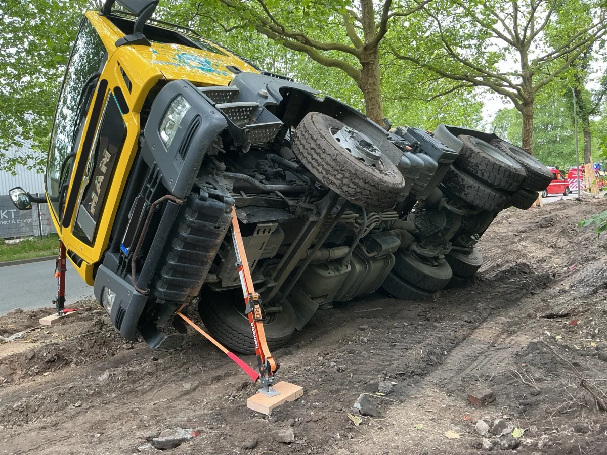 Ein gelber LKW ist auf einer schmutzigen Bodenfläche umgekippt. Bäume sind im Hintergrund zu sehen, Sicherheitsgurte stabilisieren das Fahrzeug.