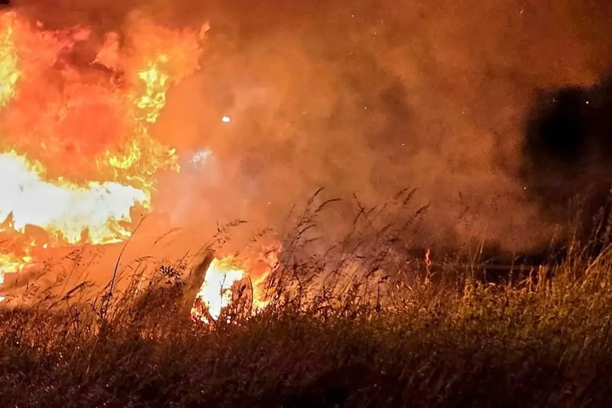 Ein intensiver Brand mit großen Flammen und starkem Rauch breitet sich in einem offenen Feld aus. Gras im Vordergrund ist ebenfalls betroffen.