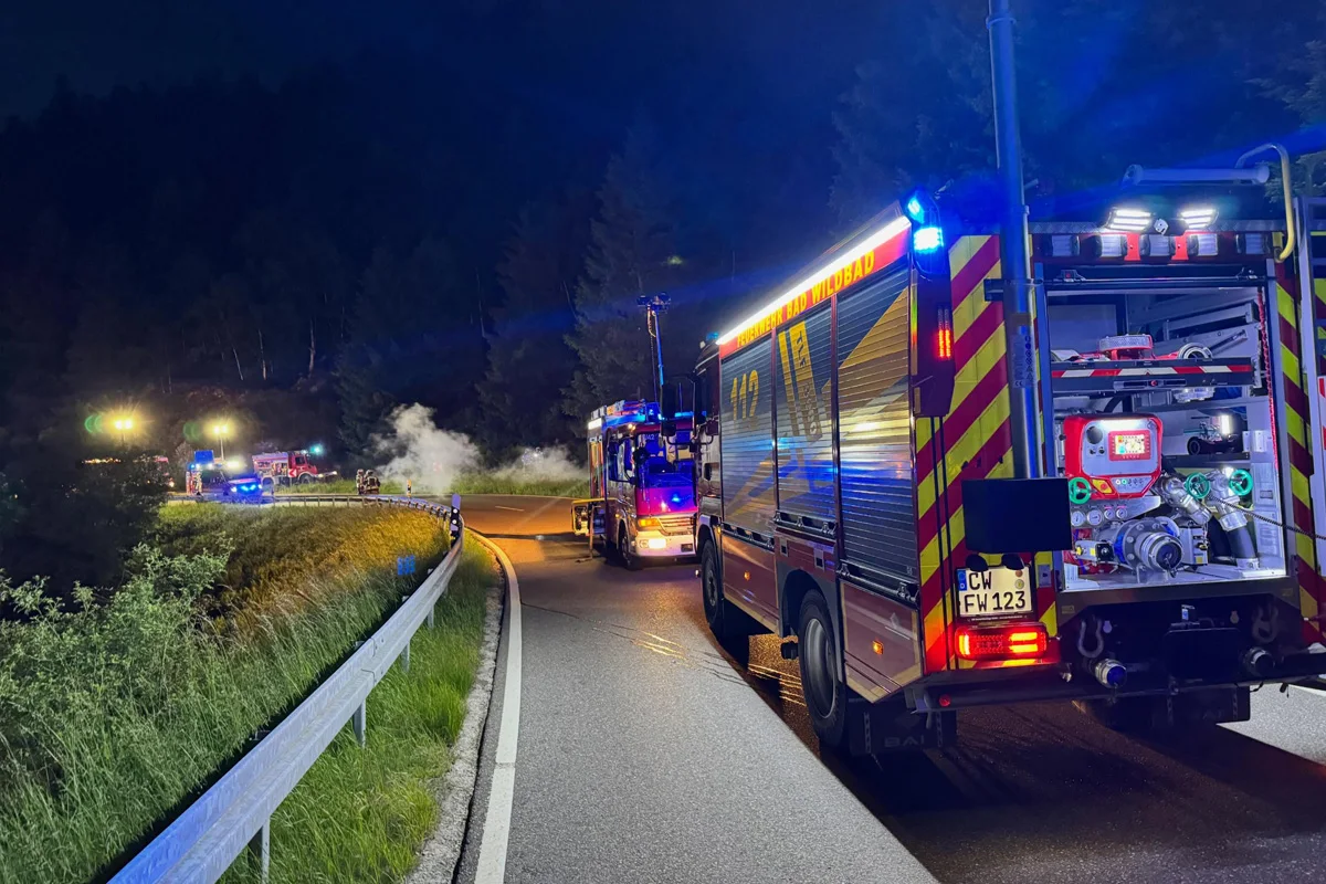 Mehrere Feuerwehrfahrzeuge stehen nachts auf einer kurvigen Landstraße, die beleuchtet ist. Rauch ist in der Ferne sichtbar.
