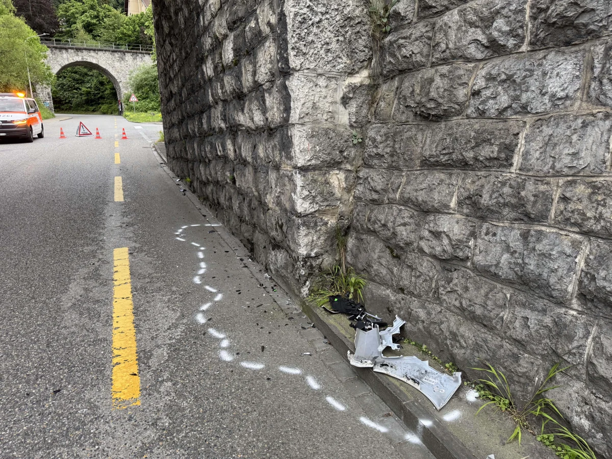 Zu sehen sind Trümmerteile eines Fahrzeugs an einer markierten Unfallstelle entlang einer grauen Tunnelmauer. Im Hintergrund sind Absperrungen und ein Fahrzeug mit Blaulicht erkennbar.