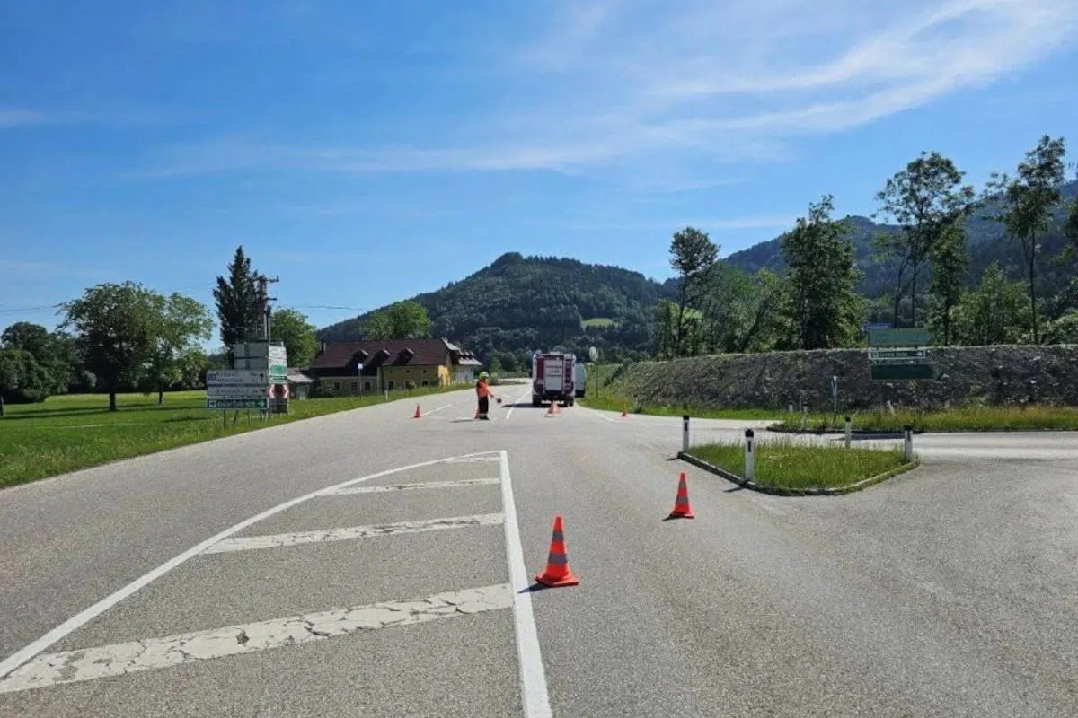 Auf einer Landstraße steht ein Feuerwehrfahrzeug, umgeben von Verkehrskegeln bei sonnigem Wetter. Im Hintergrund sind Bäume und ein Berg zu sehen.