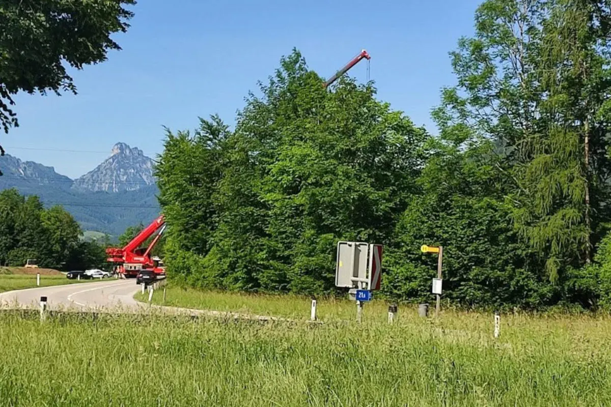 Ein roter Kranwagen steht auf einer kurvigen Landstraße im Vordergrund. Im Hintergrund sind dichte Bäume und ein Berg zu sehen.