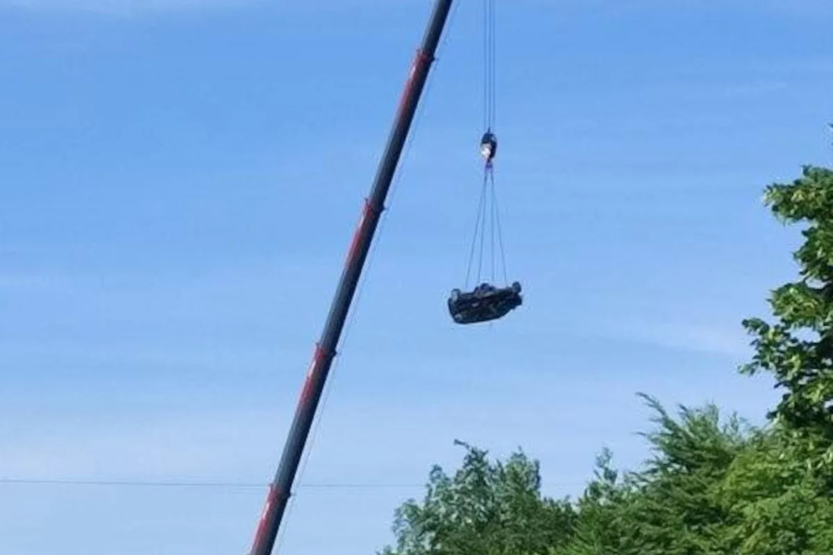Ein Kran hebt ein Auto in die Luft. Das Fahrzeug hängt mit Seilen am Kranhaken. Der Himmel ist blau und keine Personen sind sichtbar.