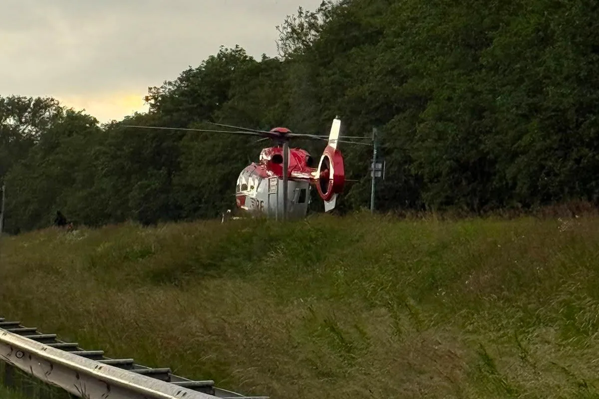 Ein rot-weißer Rettungshubschrauber steht auf einer Wiese neben einer bewaldeten Straße, bereit für einen Einsatz.