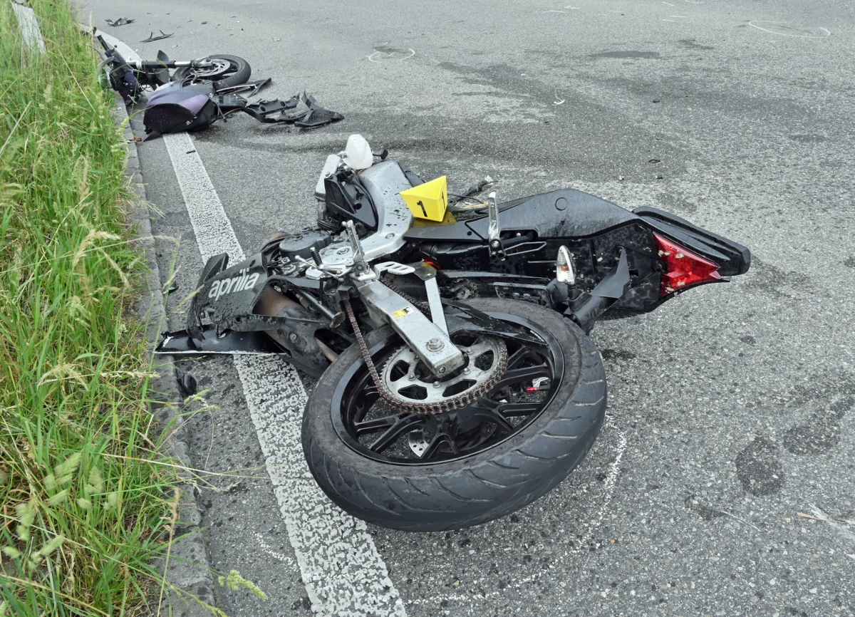 Ein beschädigtes Motorrad liegt auf der Fahrbahn, umgeben von verstreuten Trümmern, nahe der Straßenlinie mit hohem Gras.