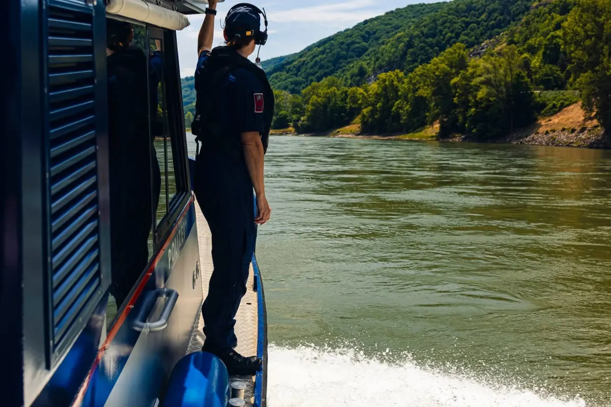 Ein Polizist steht auf dem Deck eines blauen Polizeiboots und blickt auf den Fluss. Im Hintergrund sind bewaldete Ufer zu sehen.