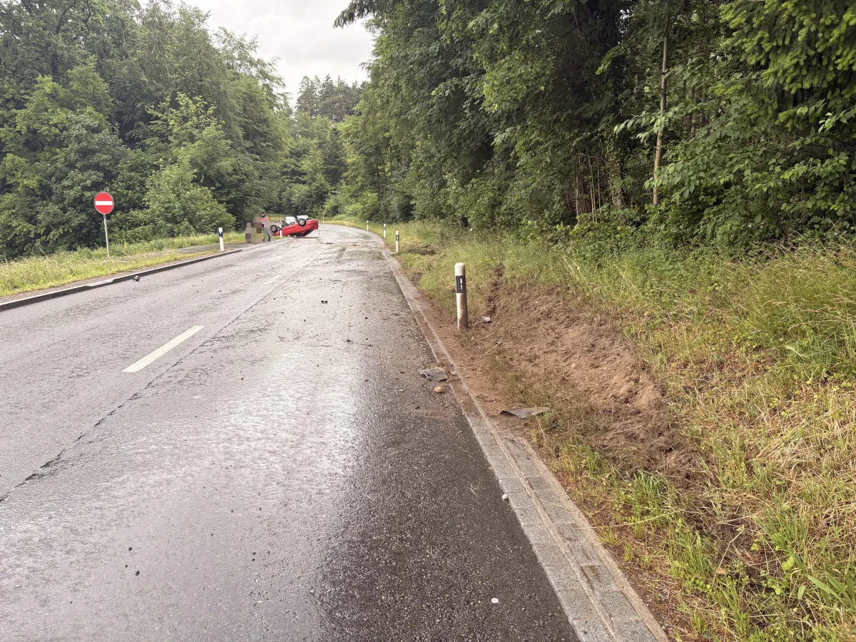 Ein rotes Auto steht am Straßenrand einer Landstraße, die durch einen Wald führt. Der Boden ist nass, was auf Regen hindeutet.