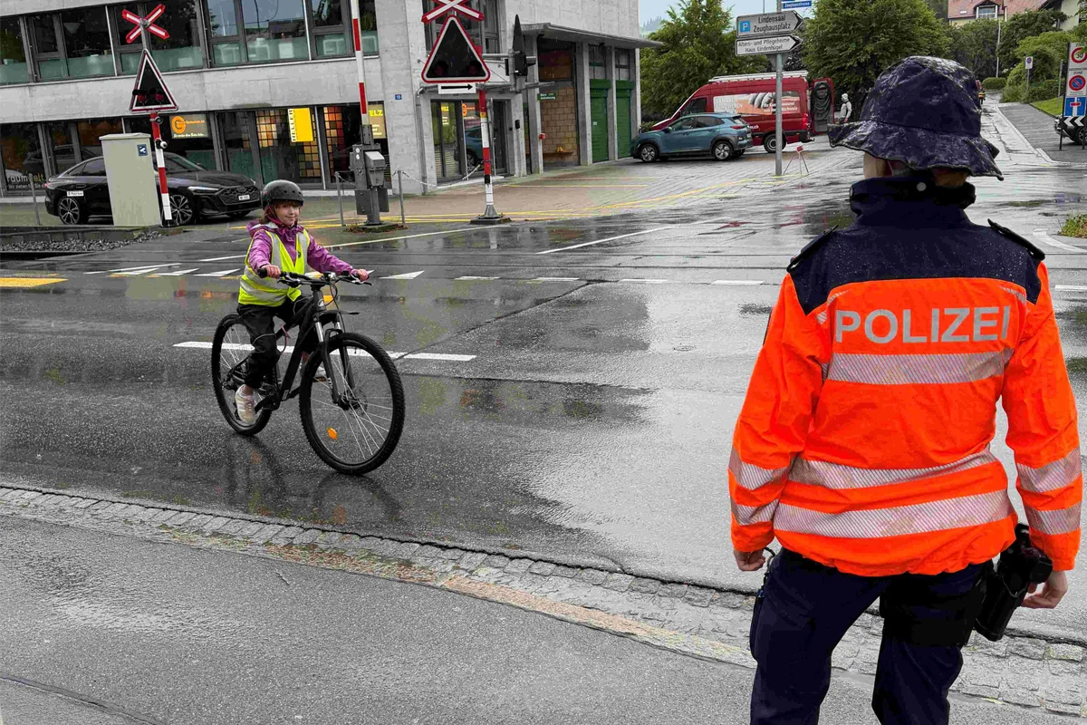 Ein Polizist in einer orangefarbenen Jacke steht an einem Bahnübergang. Ein Kind fährt in einer gelben Warnweste auf einem Fahrrad vorbei.