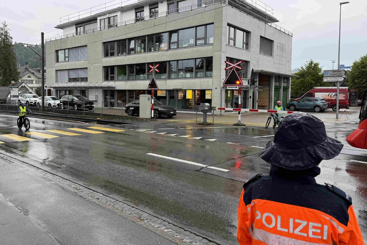 Ein Polizist in orange-blauer Uniform steht vor einem gesperrten Bahnübergang. Es regnet und die Straße ist nass.