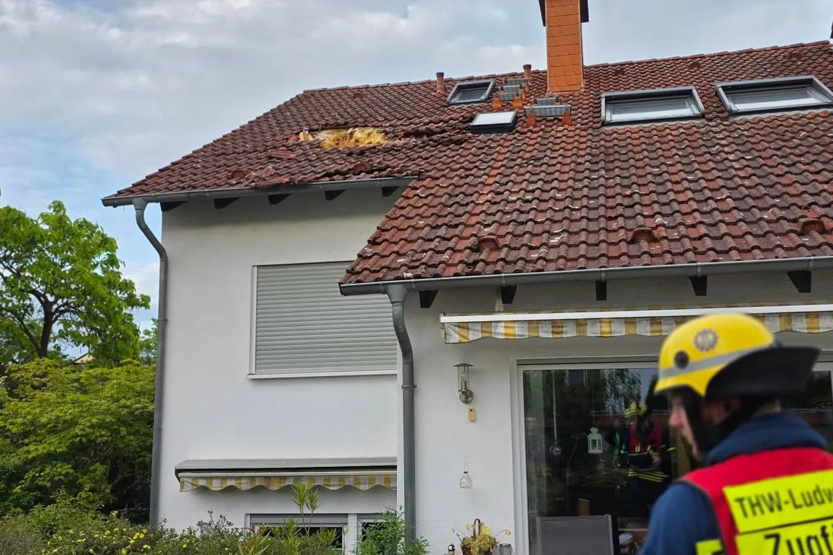 Ein Haus mit einem roten Ziegeldach zeigt sichtbare Beschädigungen durch ein Unwetter. Ein Einsatzhelfer des Technischen Hilfswerks steht im Vordergrund.