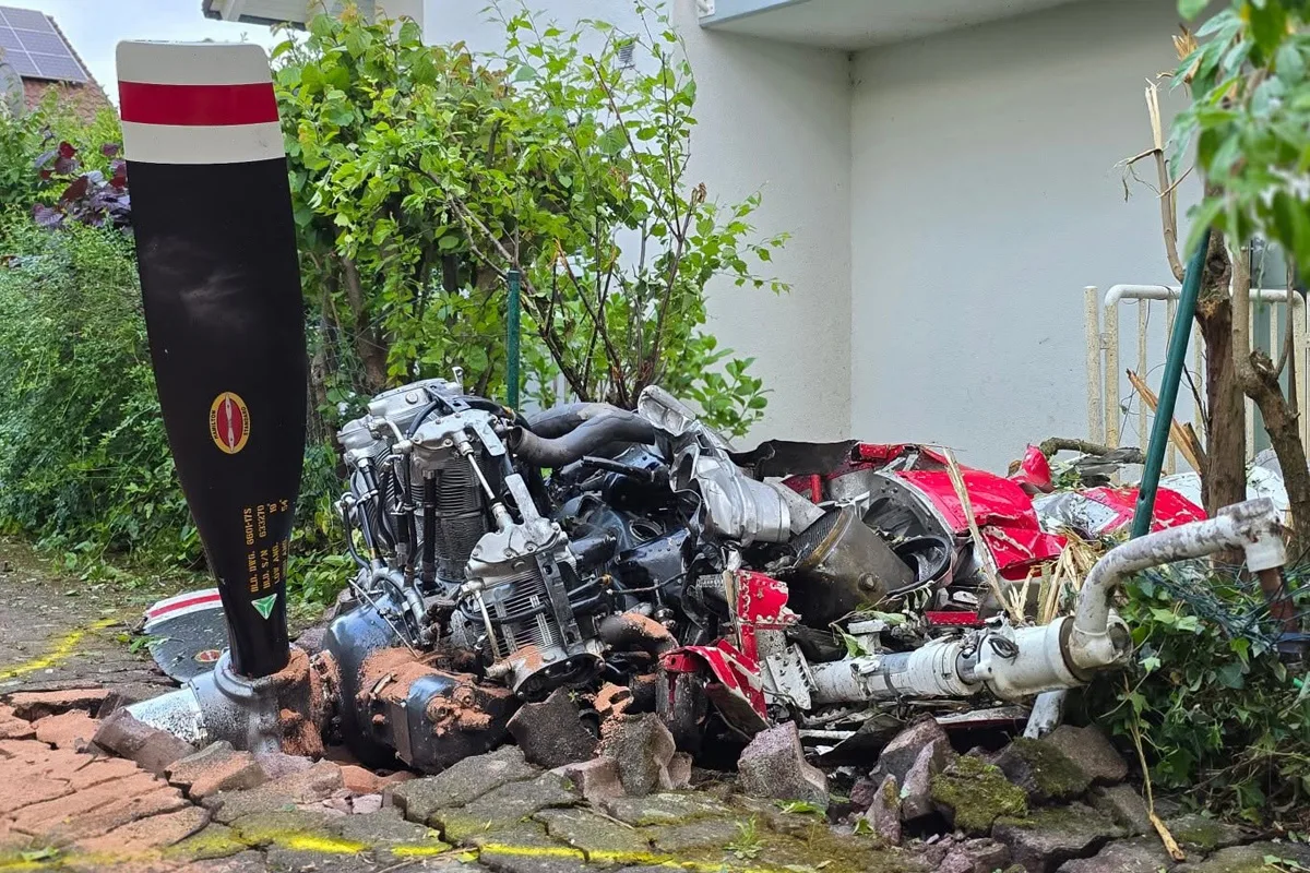 In einem Garten liegen beschädigte Hubschraubertrümmer. Ein großer Propeller ist sichtbar, umgeben von Pflanzen und beschädigtem Boden.