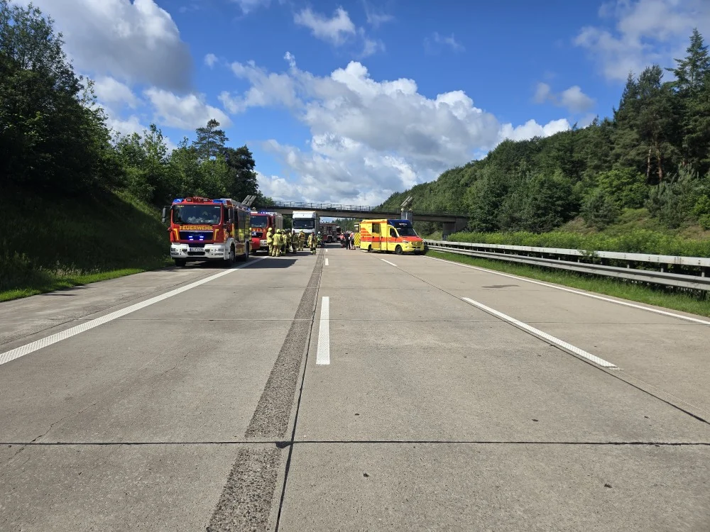 Ein Feuerwehr- und ein Rettungsfahrzeug blockieren eine Autobahn. Es sind weitere Einsatzkräfte vor Ort, der Himmel ist bewölkt, aber klar.