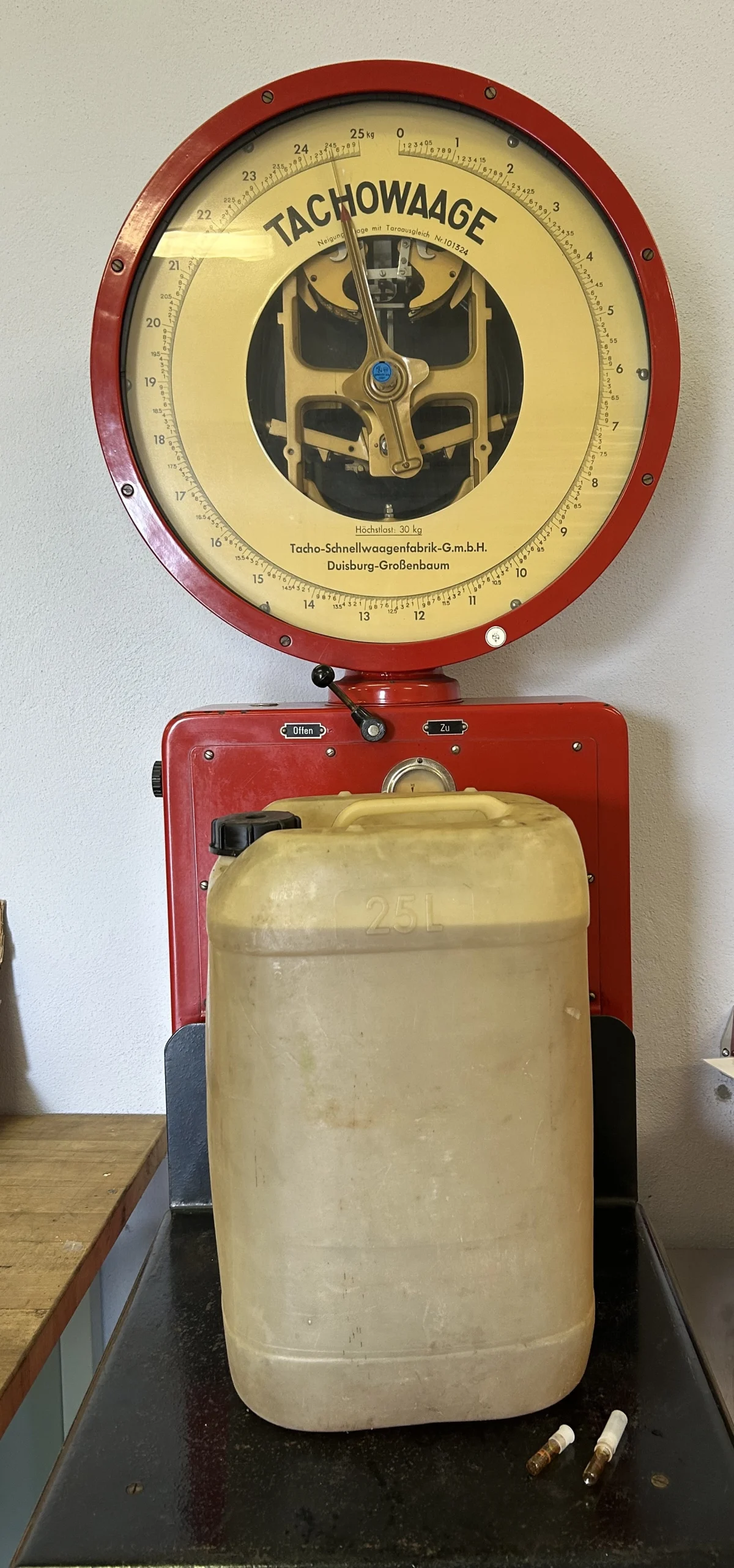 Eine antike rote Waage zeigt ihr Zifferblatt, während ein 25-Liter-Kanister darauf steht. Im Hintergrund ist eine Werkstattwand erkennbar.