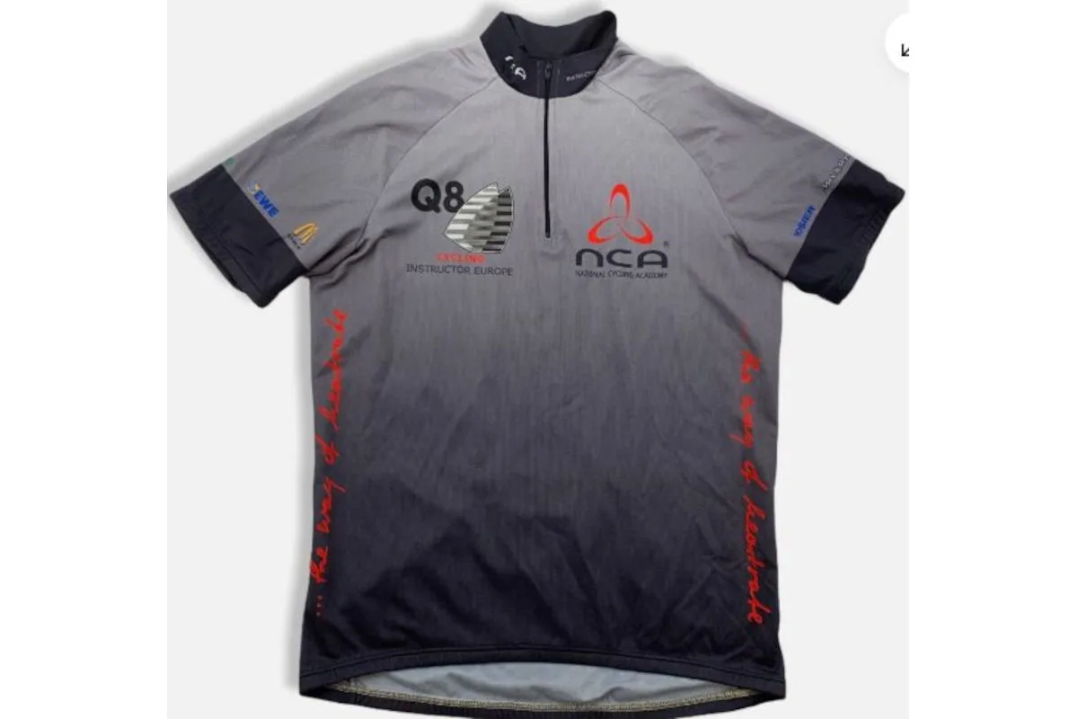 Ein graues Trikot liegt flach mit sichtbaren Logos der Marken Q8 und NCA. Der Slogan 'the way of leadership' ist aufgedruckt.