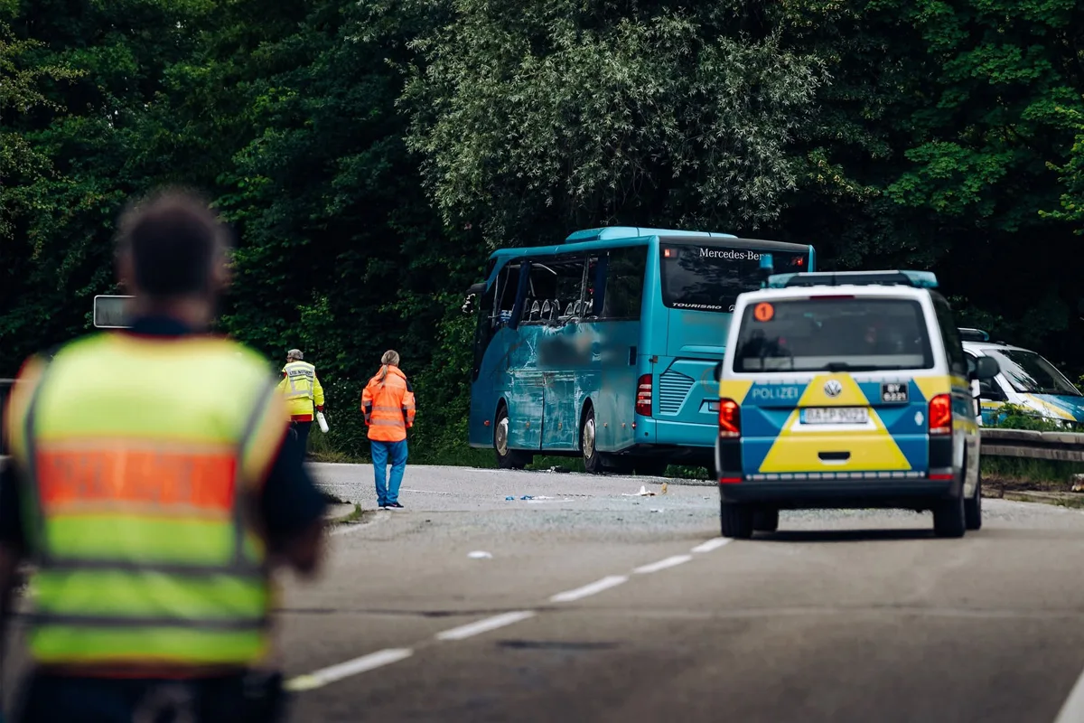 Ein blauer Reisebus zeigt deutliche Beschädigungen auf der linken Seite, während er neben einem Polizeifahrzeug auf einer Straße steht. Personen in Warnwesten befinden sich in der Umgebung.