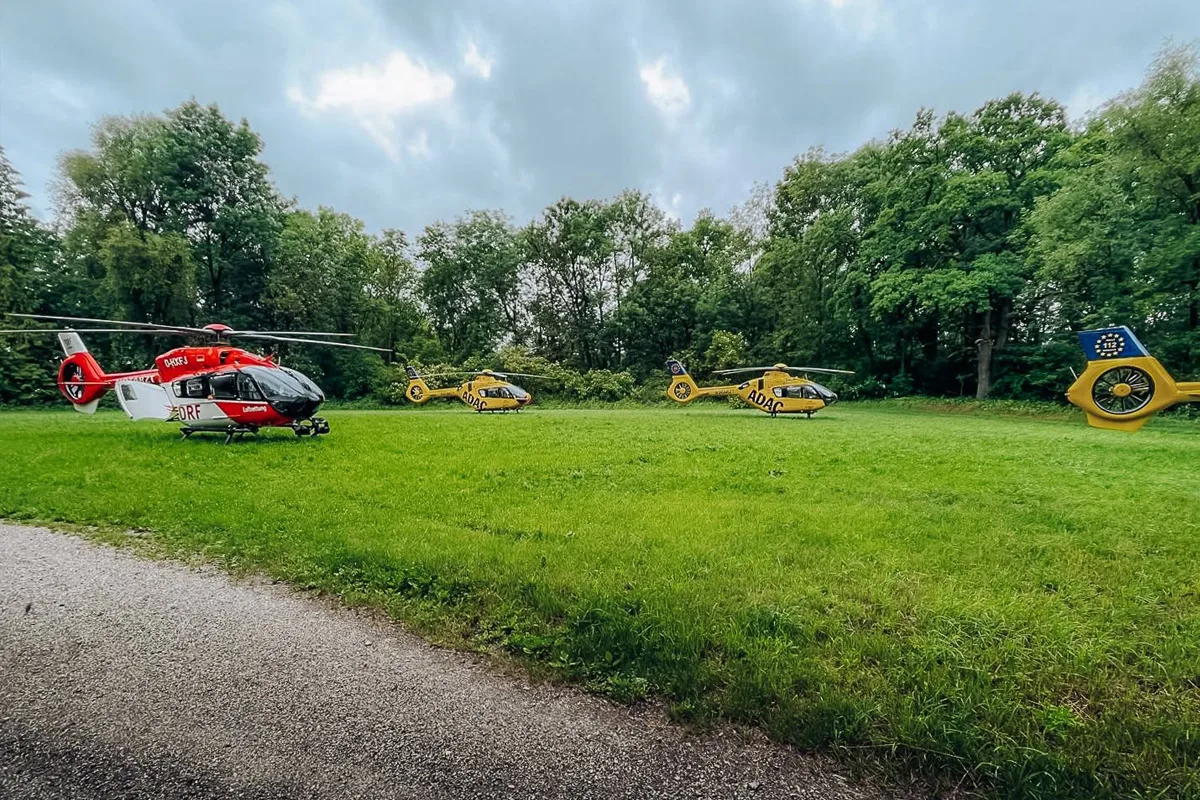 Auf einer grünen Wiese stehen drei Rettungshubschrauber, erkennbar an den Aufschriften 'ADAC'. Im Hintergrund ist ein von Bäumen gesäumter Wald zu sehen.