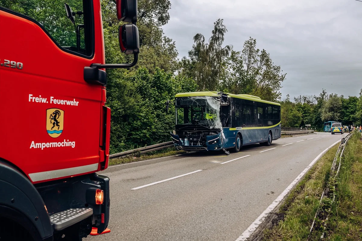 Ein Linienbus mit stark beschädigter Frontscheibe steht in Fahrtrichtung auf einer Landstraße. Ein Feuerwehrauto steht am Straßenrand.