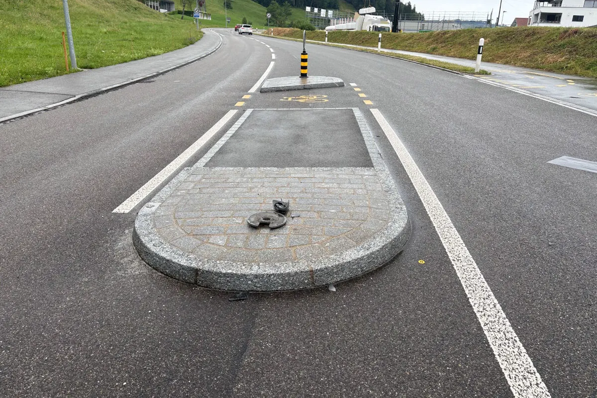 Eine Verkehrsinsel auf einer Landstraße zeigt eine abgerissene Schraube in der Mitte. Die Straße ist leer bis auf ein Fahrzeug im Hintergrund, bei bewölktem Wetter.