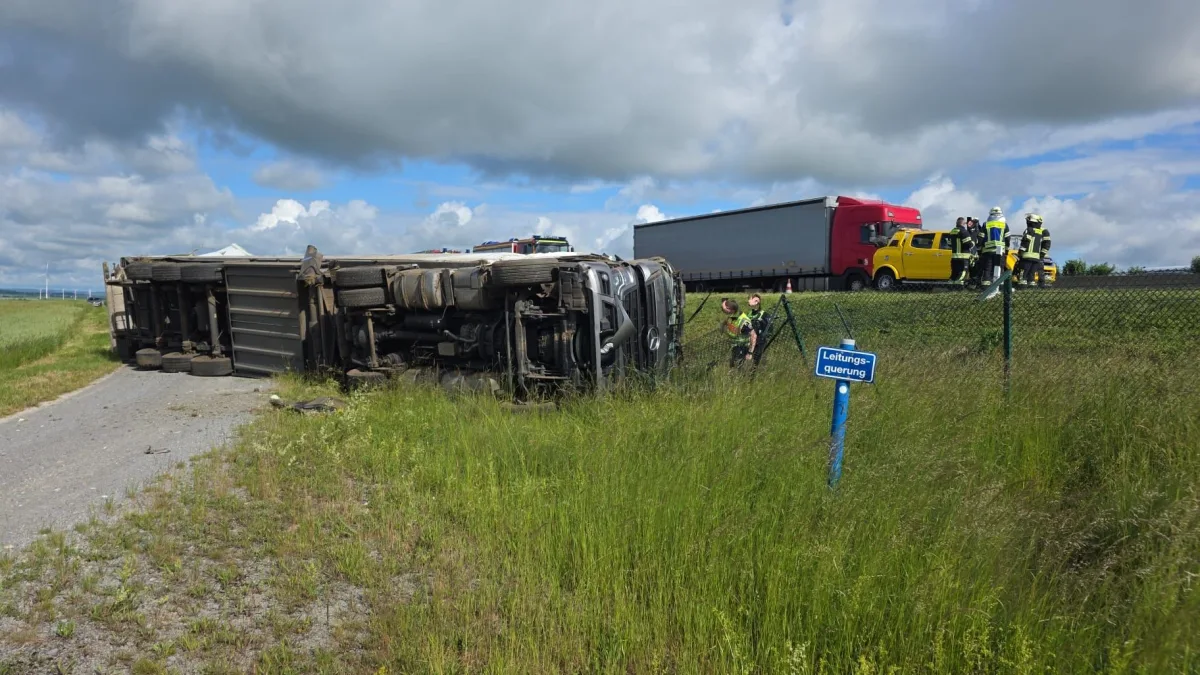 Ein umgekippter Lastwagen liegt neben einer Autobahn, während Feuerwehrleute vor Ort sind. Ein weiteres Fahrzeug ist im Hintergrund zu sehen.