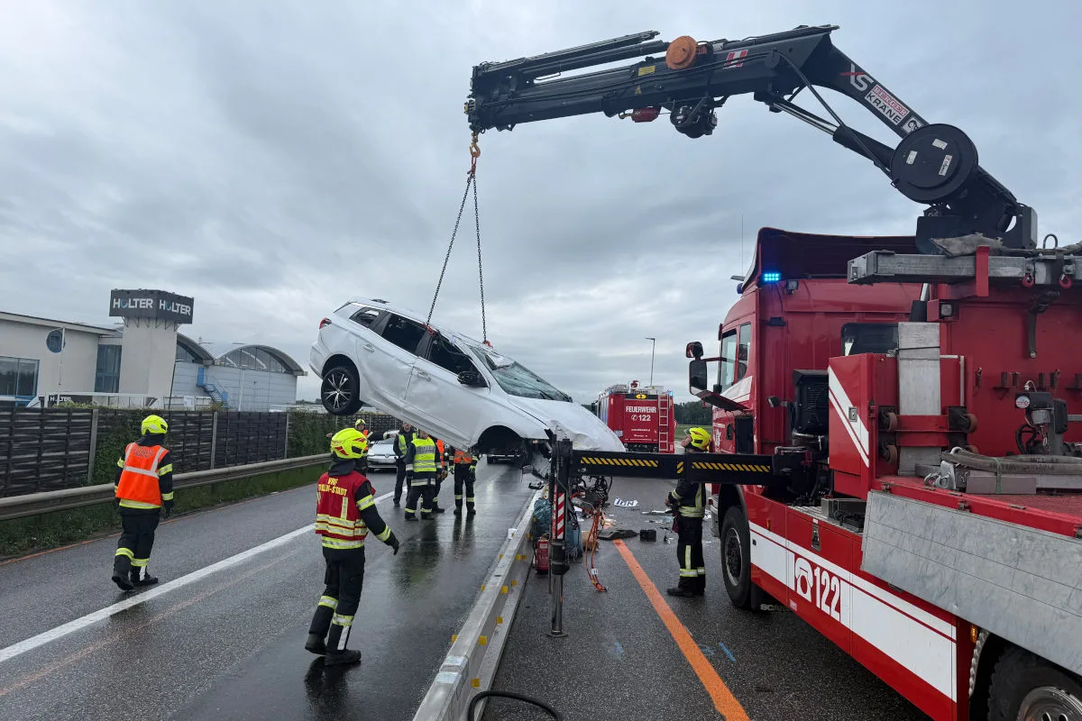 Ein Kran hebt ein beschädigtes weißes Auto von einer Autobahn. Feuerwehrleute in Schutzkleidung sind vor Ort.