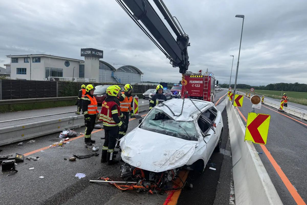 Ein zerstörtes, weißes Auto steht auf einer Autobahn, umgeben von Feuerwehrleuten in gelben Helmen und orangefarbenen Westen. Ein Feuerwehrwagen steht im Hintergrund.