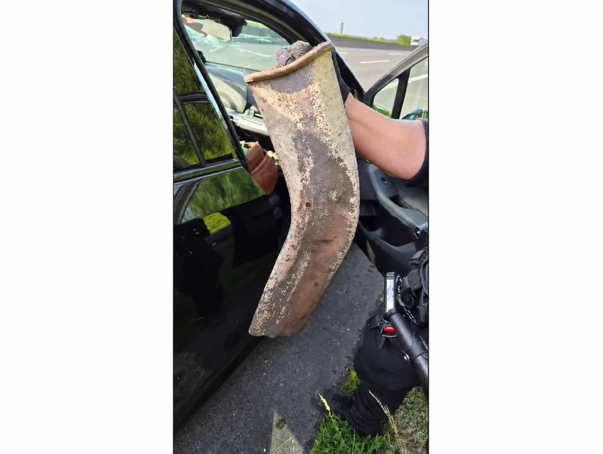 Das Bild zeigt eine Hand, die ein großes, rostiges und gebogenes Auspuffteil hält. Im Hintergrund ist ein schwarzes Auto mit geöffneter Tür zu sehen.