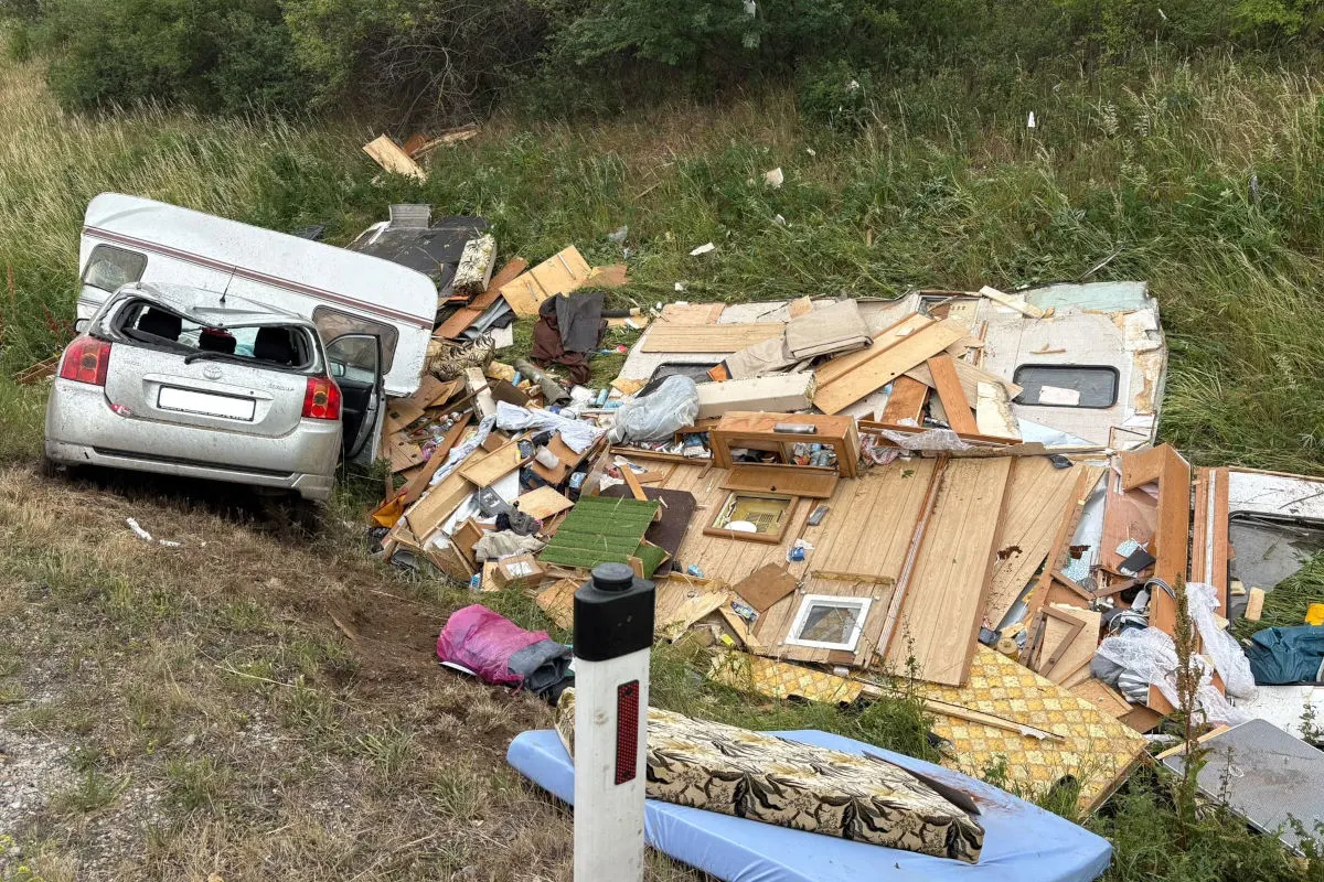 Ein verunglücktes Auto ist auf einer Böschung, der angekoppelte Wohnwagen ist zerstört und Trümmer sind verstreut. Gras wächst in der Umgebung.